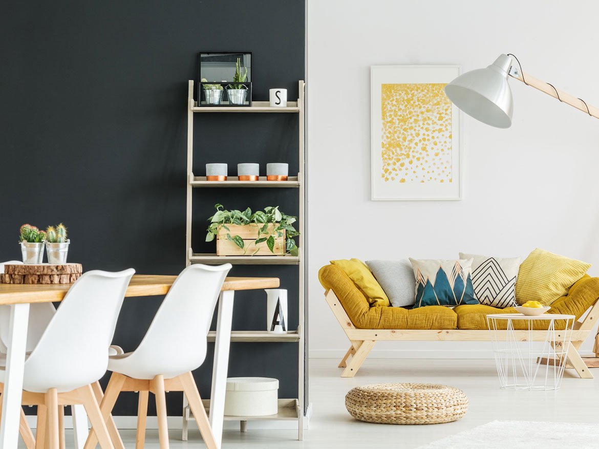 A modern living area in new homes in GTA features a yellow couch with patterned pillows, a wooden coffee table, and a white floor lamp. Adjacent is a dining area with white chairs and a wooden table. A wall-mounted shelf holding plants and decor separates the two spaces.