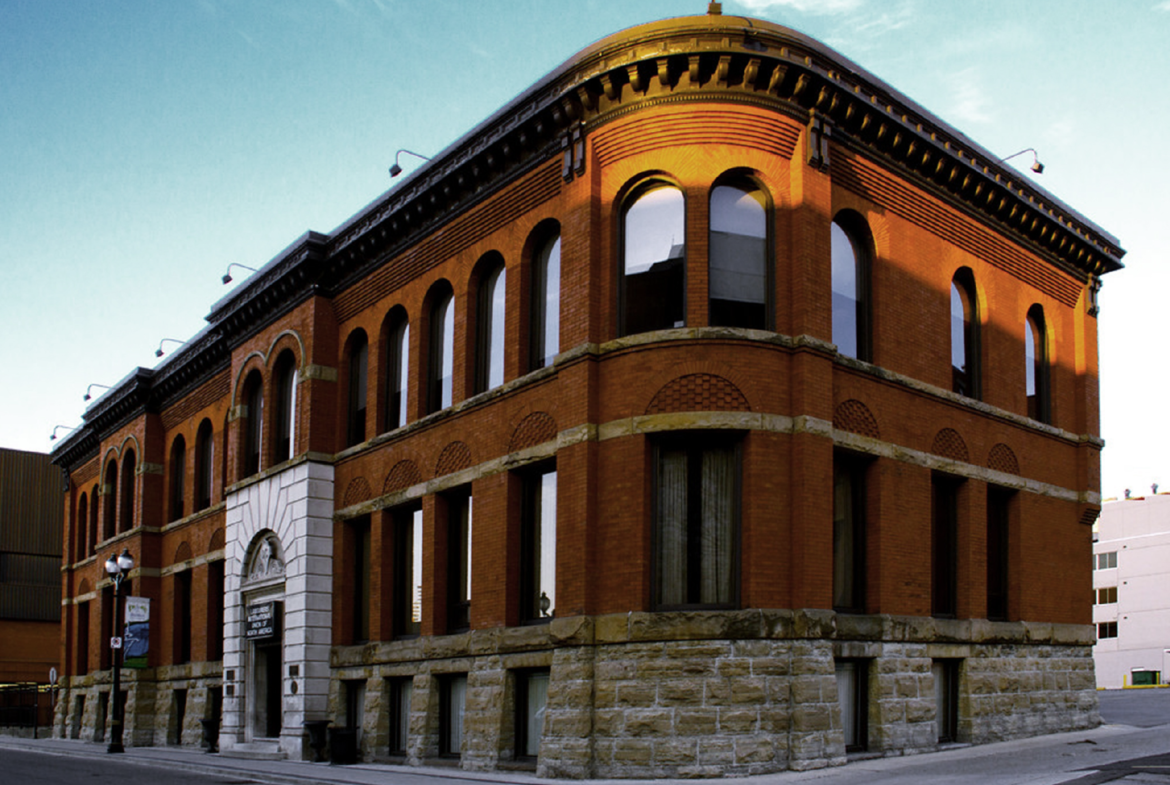 A historic, red-brick building with arched windows and a stone foundation stands on a street corner. The building features a rounded corner with a sloping roof, and the clean blue sky suggests it is a clear day. Surrounding structures, including new condos in the GTA, are modern and boxy.