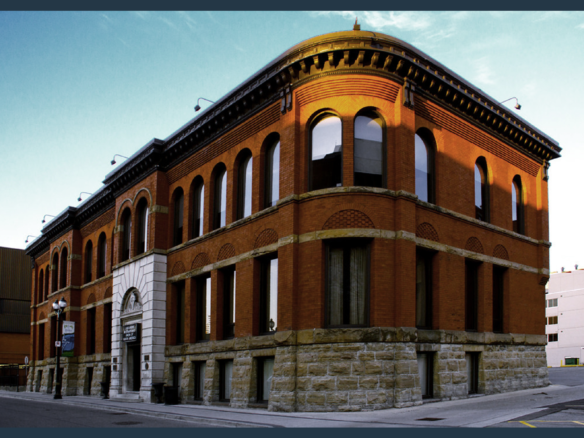 A historic, red-brick building with arched windows and a stone foundation stands on a street corner. The building features a rounded corner with a sloping roof, and the clean blue sky suggests it is a clear day. Surrounding structures, including new condos in the GTA, are modern and boxy.