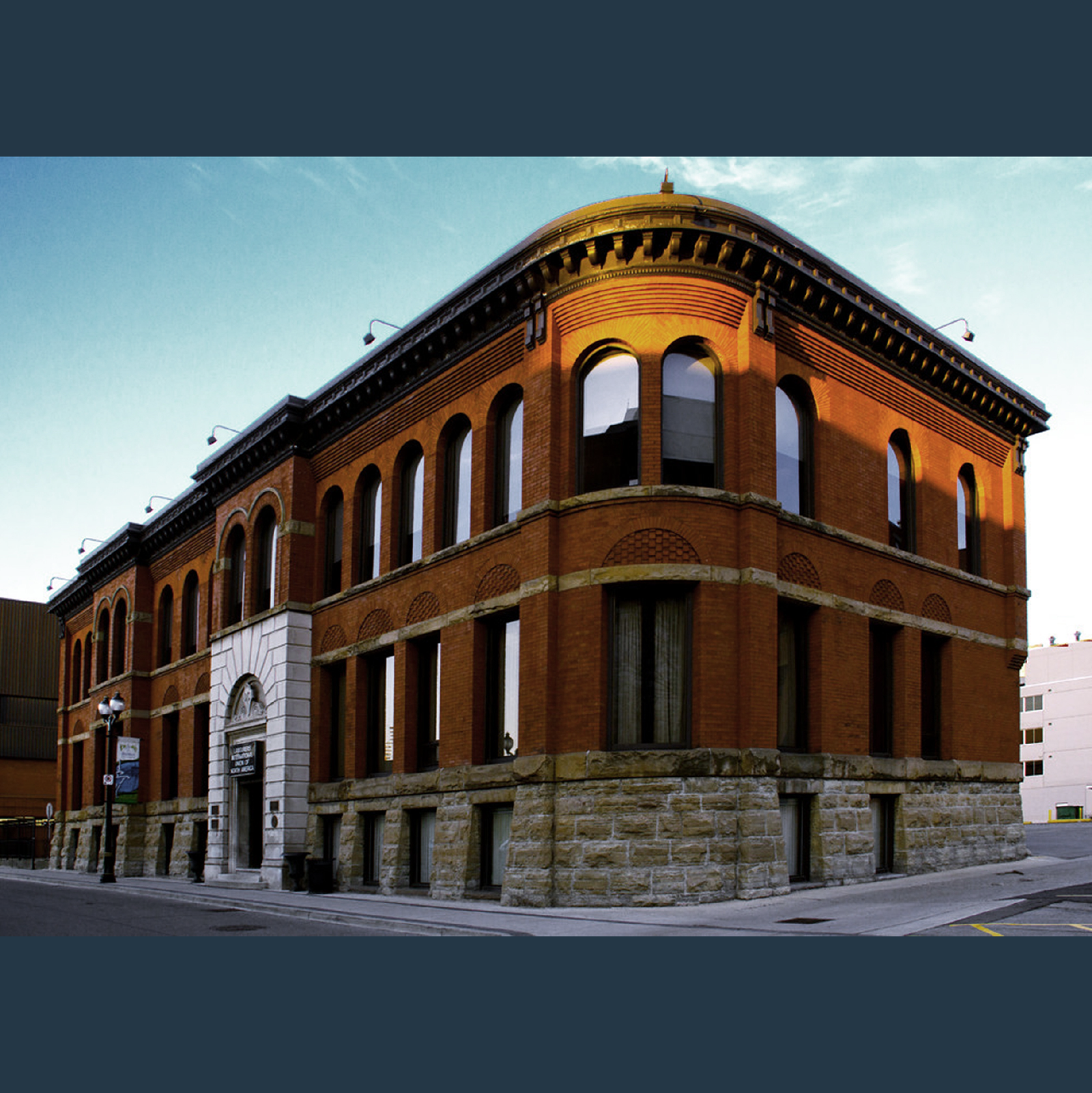 A historic, red-brick building with arched windows and a stone foundation stands on a street corner. The building features a rounded corner with a sloping roof, and the clean blue sky suggests it is a clear day. Surrounding structures, including new condos in the GTA, are modern and boxy. A historic, red-brick building with arched windows and a stone foundation stands on a street corner. The building features a rounded corner with a sloping roof, and the clean blue sky suggests it is a clear day. Surrounding structures, including new condos in the GTA, are modern and boxy.
