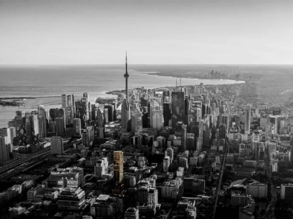 Aerial view of a sprawling cityscape with a prominent tower at its center. The city is bordered by a large body of water on the left, and new condos in Brampton stand out among the monochrome buildings, highlighted by one in yellow.