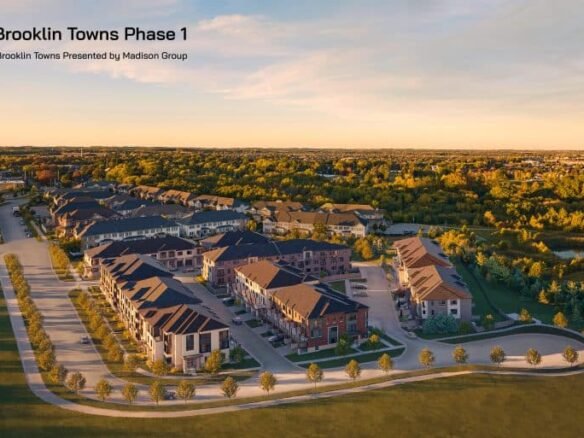 Aerial view of a suburban residential development called "Brooklin Towns Phase 1" by Madison Group. The image shows rows of townhouses with driveways and green spaces, set against a background of trees and a partly cloudy sky at sunset. Brooklin Towns promises tranquil living amidst natural beauty.