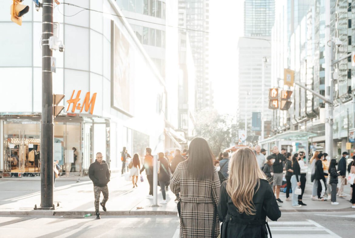 Two women, one in a plaid coat and the other in a black coat and knee-high boots, walk across a crosswalk in a bustling city street. Numerous pedestrians can be seen, and there's an H&M store in the background. Tall buildings line the street on a sunny day, hinting at new condos in GTA nearby.