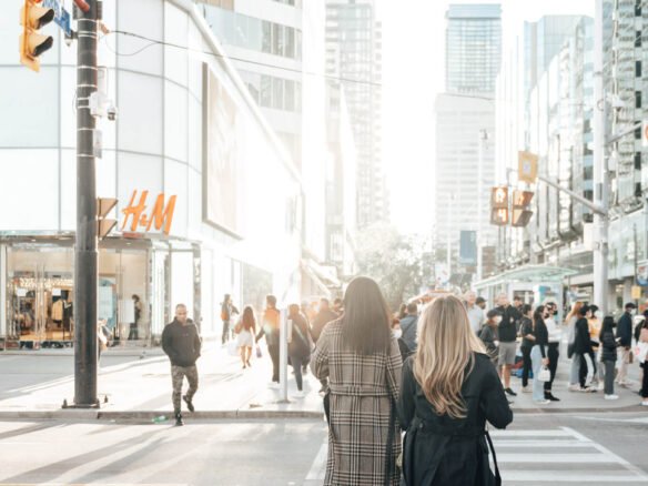 Two women, one in a plaid coat and the other in a black coat and knee-high boots, walk across a crosswalk in a bustling city street. Numerous pedestrians can be seen, and there's an H&M store in the background. Tall buildings line the street on a sunny day, hinting at new condos in GTA nearby.