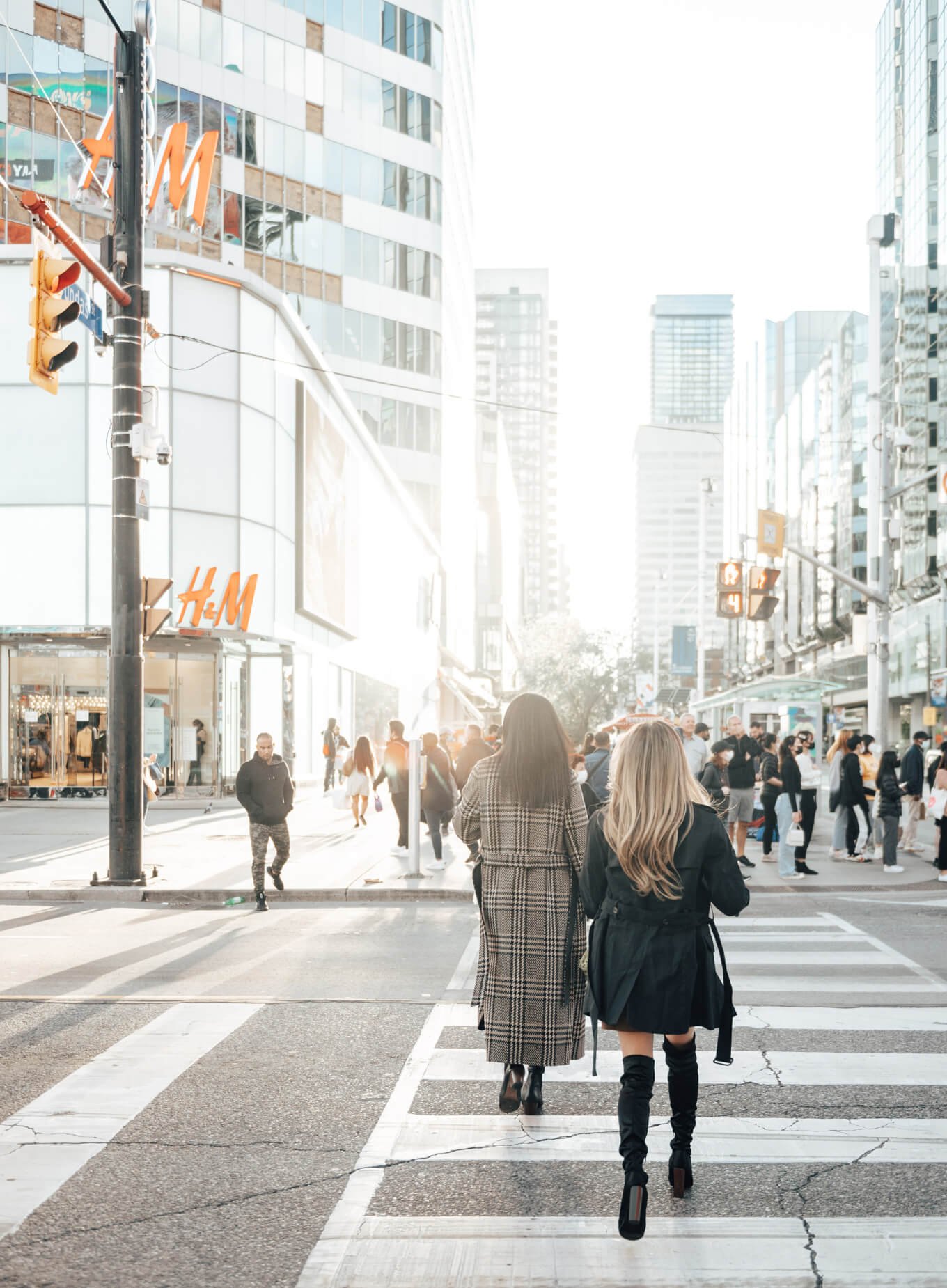Two women, one in a plaid coat and the other in a black coat and knee-high boots, walk across a crosswalk in a bustling city street. Numerous pedestrians can be seen, and there's an H&M store in the background. Tall buildings line the street on a sunny day, hinting at new condos in GTA nearby.