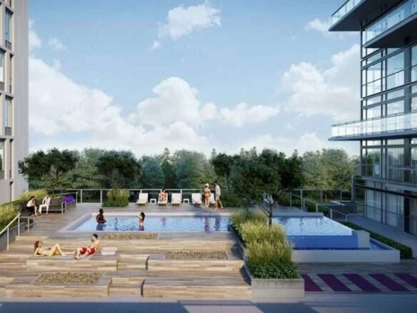 A modern rooftop pool area with two pools surrounded by lounge chairs, plants, and people relaxing. The area is bordered by tall glass buildings and overlooks a green, tree-filled landscape beneath a partly cloudy sky—an oasis that new condos in the GTA can offer.