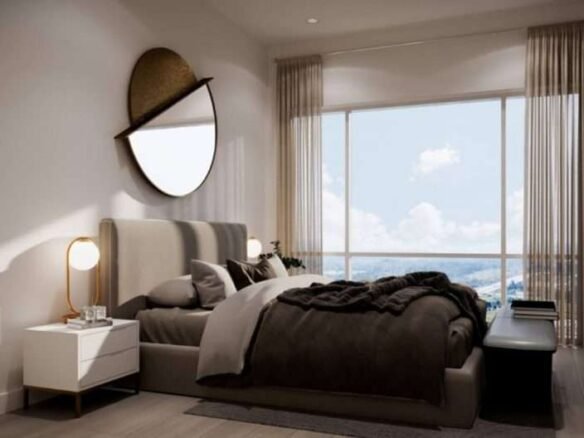 A modern bedroom in new homes in GTA featuring a large bed with dark grey bedding, a nightstand with a round lamp, and a round mirror on the wall. Light curtains partially cover a large window, allowing natural light to illuminate the room. A black bench sits at the foot of the bed.