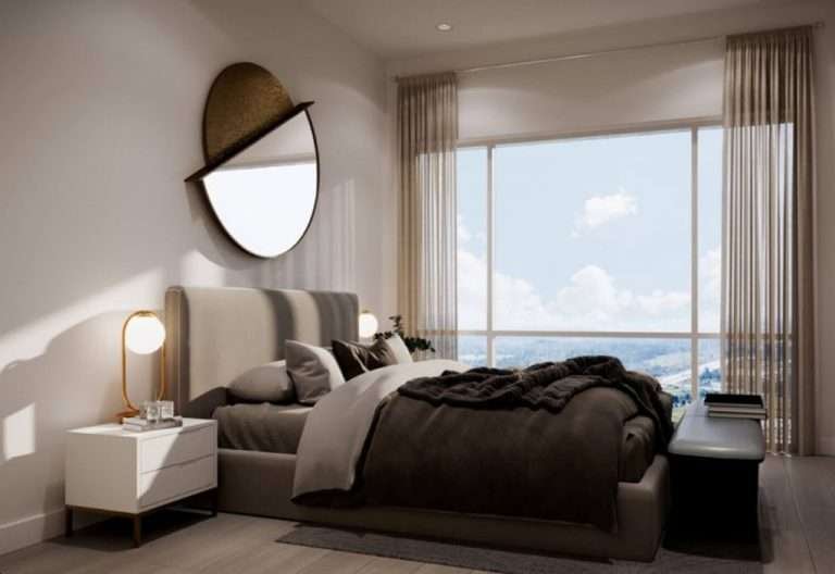 A modern bedroom in new homes in GTA featuring a large bed with dark grey bedding, a nightstand with a round lamp, and a round mirror on the wall. Light curtains partially cover a large window, allowing natural light to illuminate the room. A black bench sits at the foot of the bed.