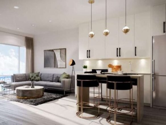 Modern open-concept living room and kitchen area with sleek white cabinetry, stainless steel appliances, and pendant lights. The living area has a gray sectional sofa, round coffee table, floor lamp, and large window with a view. New condos in GTA feature stylish rugs and wall art to complete the look.