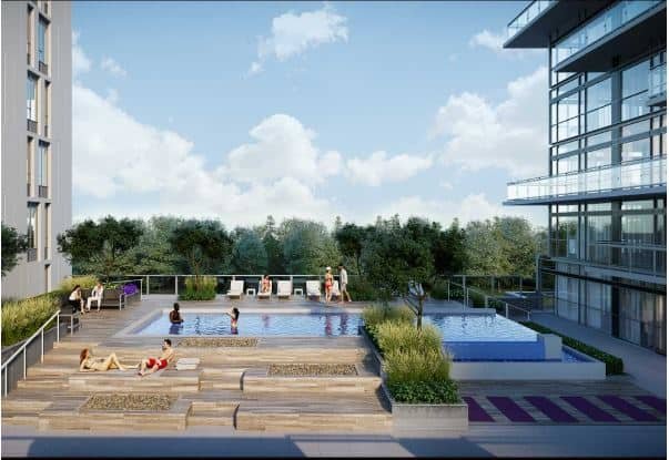 A modern outdoor pool area with two rectangular pools is surrounded by lush greenery and adjacent to new homes. People are lounging and socializing by the water, enjoying the wooden decking and planters. The area, next to a multi-story glass building, basks under a clear sky with a few clouds.