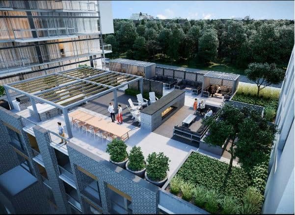 A rooftop terrace features lounge areas with outdoor furniture, a bar with stools, and a pergola providing shade. In the background, lush greenery and a body of water are visible. People enjoy the space, which is surrounded by modern buildings showcasing new condos in GTA.