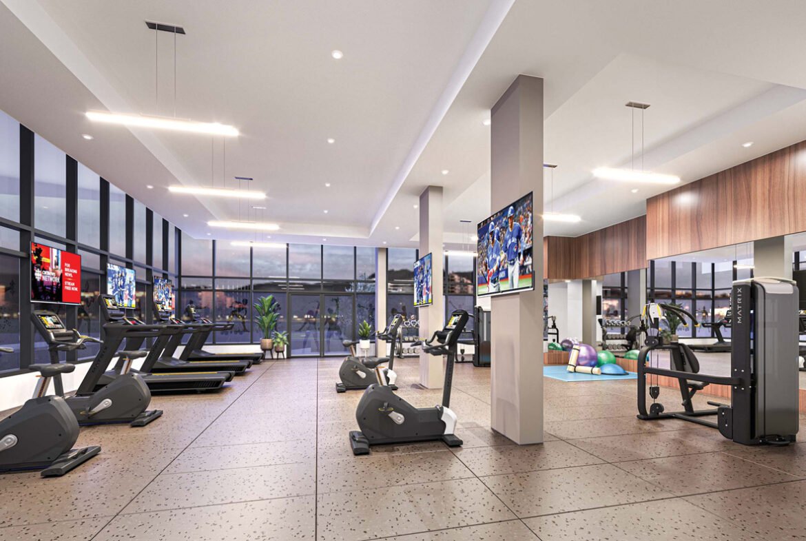 Interior of a modern gym featuring treadmills, exercise bikes, weight machines, and fitness balls. TVs mounted on walls and large windows in a well-lit space.
