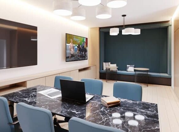 A modern conference room at Motto Condos features a polished black marble table, blue chairs, a laptop, notebooks, and a pen on the table. The room boasts a large wall-mounted TV, minimalist lighting fixtures, wood paneling, and a cozy seating nook with cushions in the background.