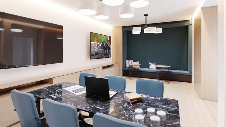A modern conference room at Motto Condos features a polished black marble table, blue chairs, a laptop, notebooks, and a pen on the table. The room boasts a large wall-mounted TV, minimalist lighting fixtures, wood paneling, and a cozy seating nook with cushions in the background.