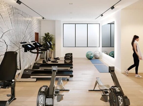 A modern gym at Motto Condos features treadmills, rowing machines, and exercise bikes lined up against a wall with a large tree ring mural. A woman stands near exercise balls and yoga mats, with bright windows bringing in natural light in the background—an ideal real estate investment for fitness enthusiasts.