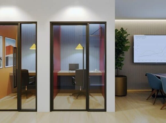 A modern office space at Motto Condos features two glass-walled private rooms with single desks and chairs. The main area boasts a conference table surrounded by blue chairs, a large plant, and a wall-mounted screen displaying charts. Artwork adorns the walls of the private rooms, enhancing the sleek design.
