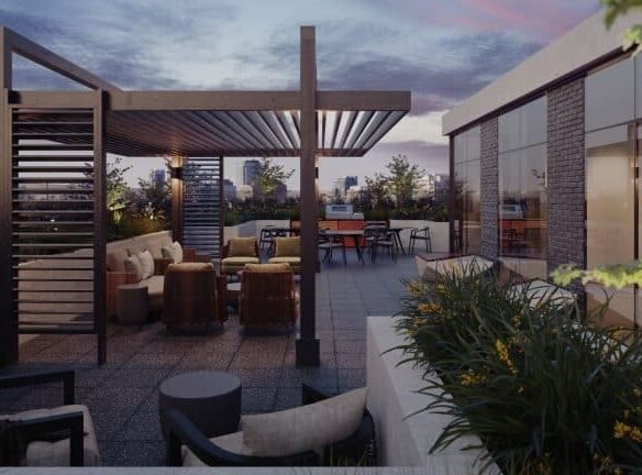 A modern rooftop terrace at dusk at Motto Condos features cushioned seating, a wooden pergola, a BBQ area, and lush potted plants. The city skyline is visible in the background under a colorful sky. The space is illuminated by subtle ambient lighting, creating an inviting atmosphere.