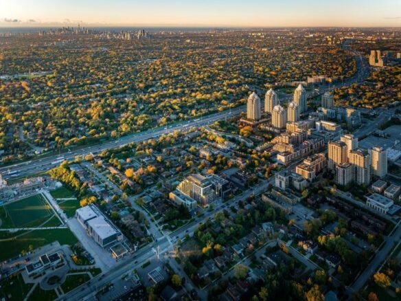 Aerial view of a suburban area at sunset featuring new condos, residential buildings, green spaces, and a main road, with a city skyline in the distance.