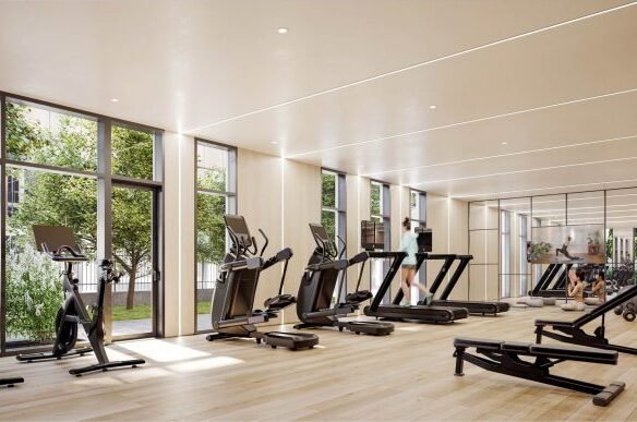 A spacious, modern gym at Six99 Condos features large windows allowing natural light to flood in. The facility includes various cardio equipment such as treadmills and stationary bikes, along with free weights and weight machines spread throughout the wooden floor area.
