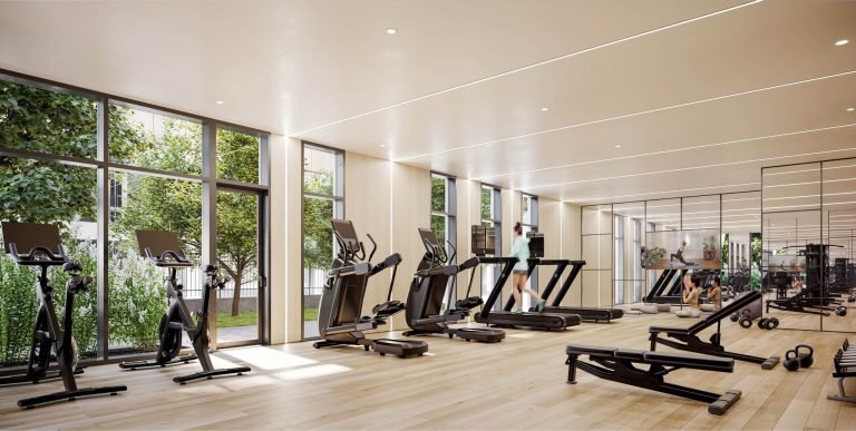 A spacious, modern gym at Six99 Condos features large windows allowing natural light to flood in. The facility includes various cardio equipment such as treadmills and stationary bikes, along with free weights and weight machines spread throughout the wooden floor area.