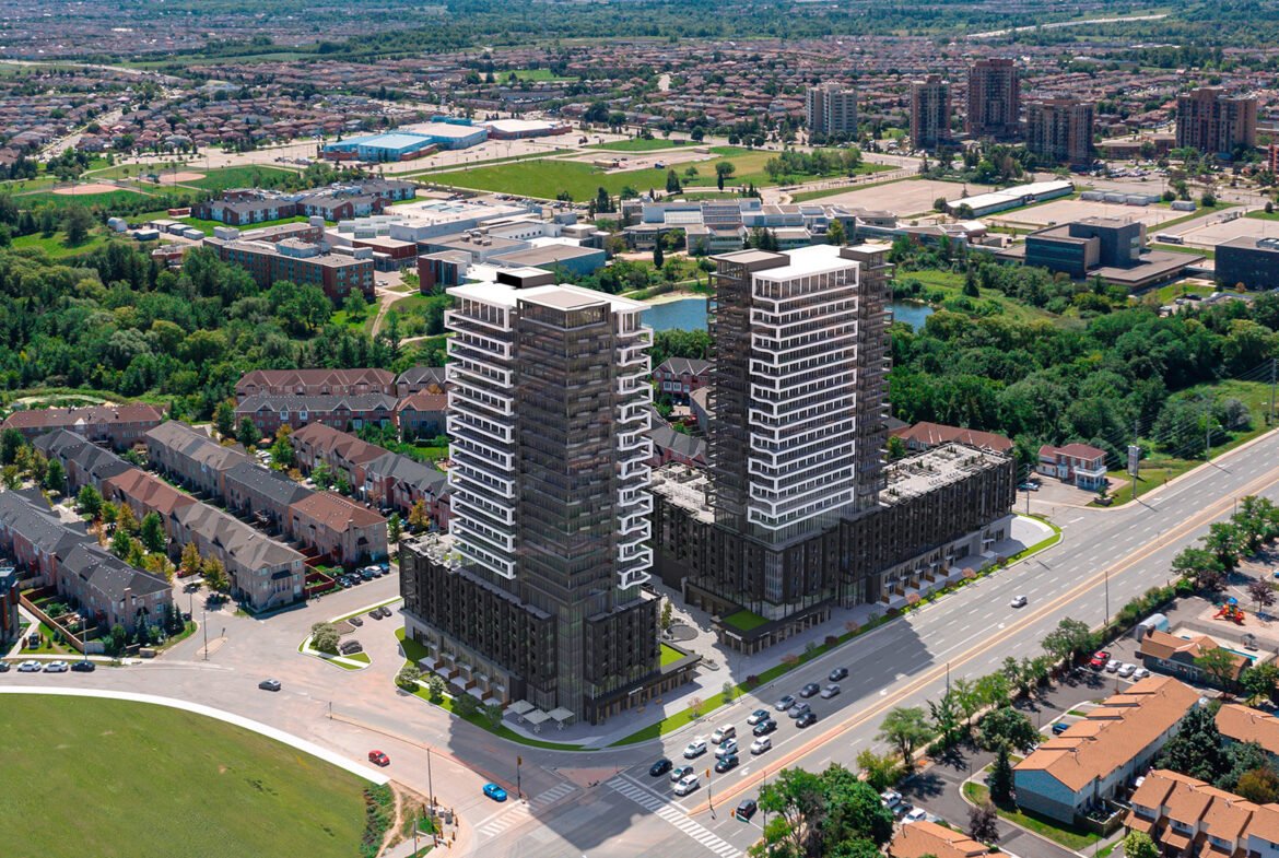 Aerial view of two tall residential towers under New condos construction beside a busy highway, surrounded by a suburban landscape with houses, smaller buildings, and patches of greenery.