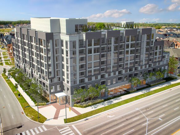 A modern, multi-story apartment building with large windows stands surrounded by trees on a street corner. The area offers a suburban feel with houses in the background and multiple lanes on the roadway. Explore new homes in this vibrant setting, perfect for those seeking contemporary living spaces.