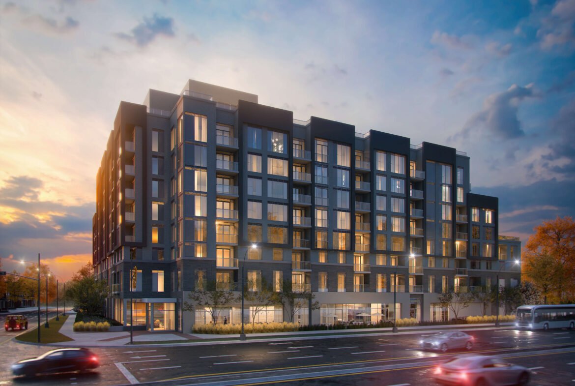 A modern apartment building with large windows glows warmly at dusk. The sky, a blend of blue and orange, sets the stage as cars blur past on the street. Trees frame this striking structure on the right, offering a glimpse into the future of new homes in Toronto.