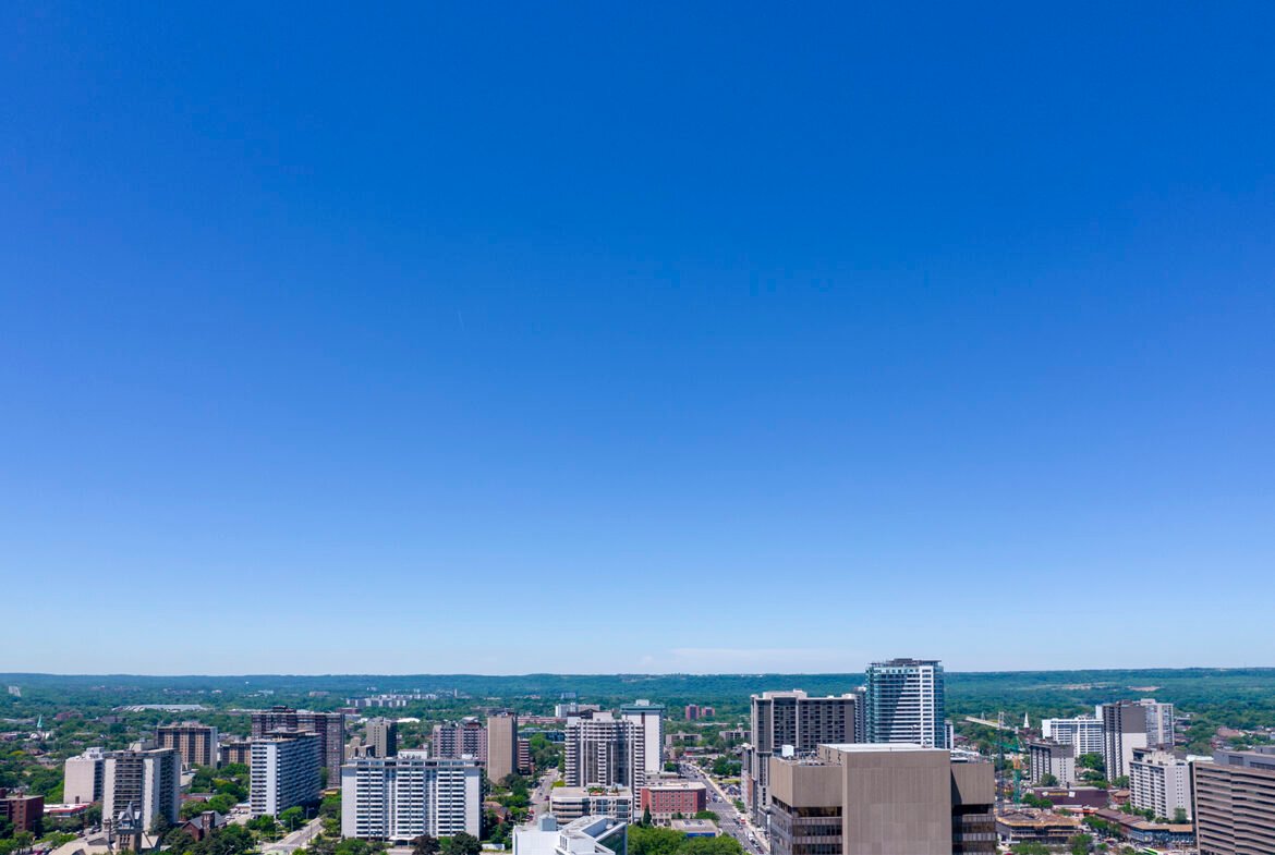 A cityscape featuring numerous high-rise buildings and skyscrapers, set against a clear blue sky. The horizon reveals green, hilly terrain in the background, blending urban and natural landscapes with new condos in GTA offering fresh perspectives on modern living.
