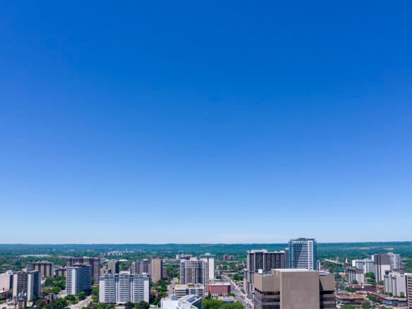 A cityscape featuring numerous high-rise buildings and skyscrapers, set against a clear blue sky. The horizon reveals green, hilly terrain in the background, blending urban and natural landscapes with new condos in GTA offering fresh perspectives on modern living.
