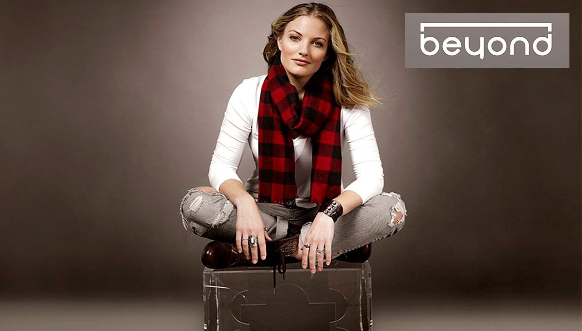A person with long hair sits cross-legged on a transparent chair, wearing a white shirt, ripped jeans, and a red and black checkered scarf. They have a confident expression. The words "Beyond Condos" appear in the upper right corner of the image.
