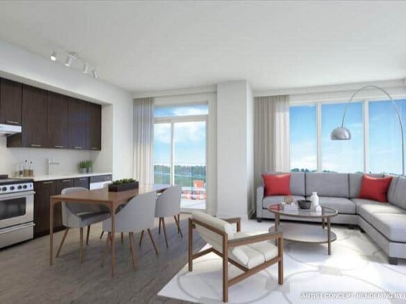 A modern, well-lit living room and kitchen area in one of the new condos in GTA features large windows providing natural light. The space boasts dark wood kitchen cabinets, a dining table with four chairs, a gray L-shaped sofa with red cushions, a round coffee table, and a standing lamp.