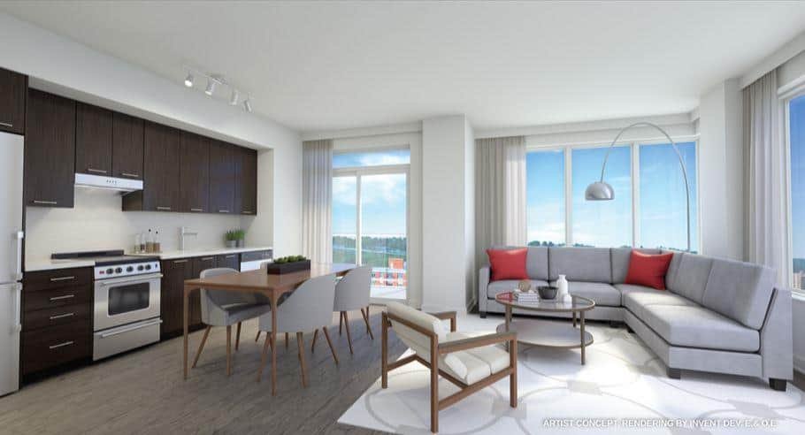 A modern, well-lit living room and kitchen area in one of the new condos in GTA features large windows providing natural light. The space boasts dark wood kitchen cabinets, a dining table with four chairs, a gray L-shaped sofa with red cushions, a round coffee table, and a standing lamp.