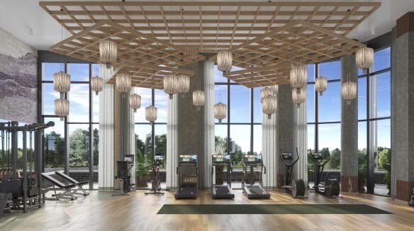 Modern gym interior with high ceilings, large windows overlooking greenery, wooden floors, and an array of exercise equipment accented by hanging lantern-style lights and a central wooden beam ceiling structure. Ideal