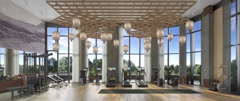 Modern gym interior with high ceilings, large windows overlooking greenery, wooden floors, and an array of exercise equipment accented by hanging lantern-style lights and a central wooden beam ceiling structure. Ideal