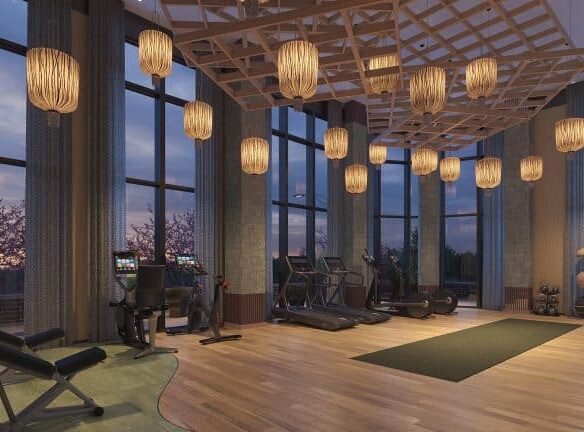 Modern gym in New Homes in GTA with large windows overlooking a sunset, featuring treadmills, elliptical machines, and hanging wooden light fixtures.