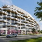 Modern wavy-designed apartment building with spacious balconies, surrounded by pink flowering trees, a lush green lawn, and a clear blue sky, situated beside a quiet street. Ideal for those looking for Pre