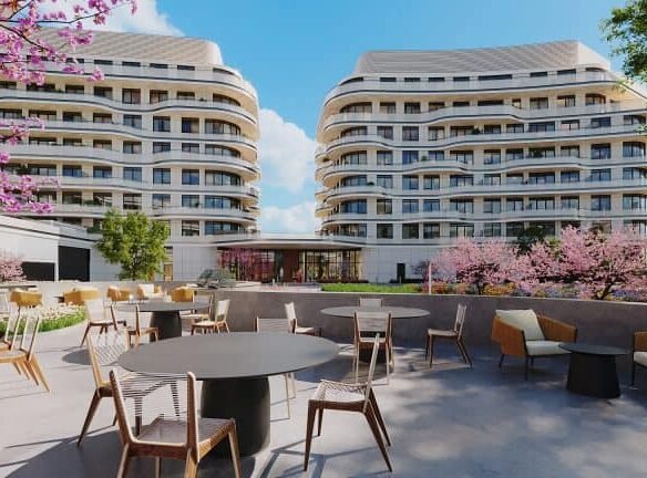 Modern outdoor café space with tables and chairs in a courtyard, framed by blossoming pink trees, showcasing New condos with curved, contemporary architecture in the background under a clear blue sky.
