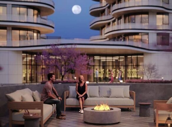 A man and woman sit around a lit fire pit on a rooftop patio with modern buildings and a full moon in the background. The scene is set during twilight, with warm ambient lighting from the surrounding buildings and blooming lily trees adding a touch of color to their serene crosstown escape.
