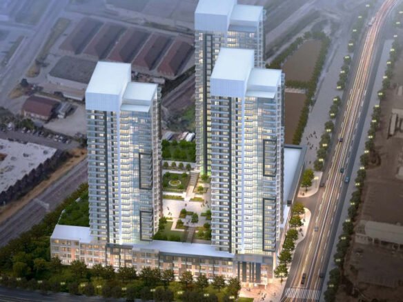 Aerial view of a modern, high-rise residential complex featuring three tall buildings with illuminated windows. The towers are connected by a green rooftop with landscaped gardens. These new condos in GTA are situated by a well-lit, bustling road lined with trees.