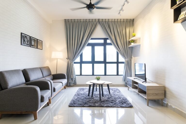 Experience modern living in one of the stunning new homes in Collingwood. The sleek living room features gray sofas, a round coffee table on a matching rug, and a flat-screen TV on a wooden stand. Large windows with blue curtains usher in natural light, highlighting minimalist decor on white walls.