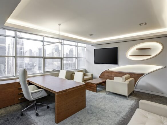Modern office with wooden desk, white swivel chair, and cream-colored seating area. Large windows offer a city skyline view, reminiscent of the sleek condos in the Dufferin Region. Walls are white with minimalist shelving and a wall-mounted TV. Natural light fills the space.