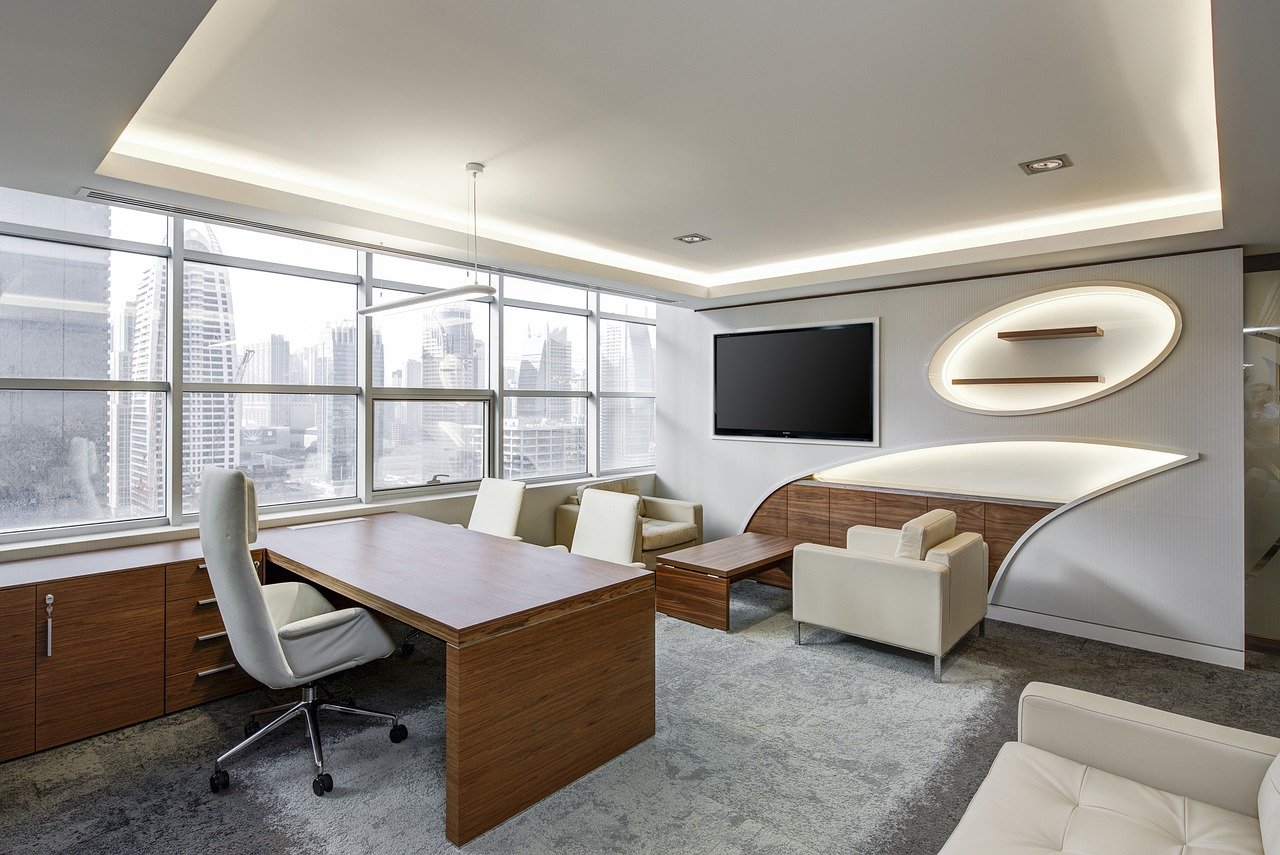 Modern office with wooden desk, white swivel chair, and cream-colored seating area. Large windows offer a city skyline view, reminiscent of the sleek condos in the Dufferin Region. Walls are white with minimalist shelving and a wall-mounted TV. Natural light fills the space.