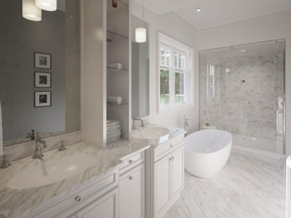 A modern bathroom in one of the new condos in GTA features a white marble floor, a double sink vanity with marble countertops, stacked towels, a freestanding oval bathtub, a glass-enclosed shower, and large windows letting in natural light.
