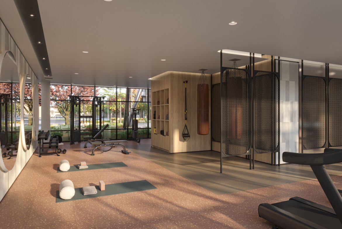 A modern gym interior at Mile & Creek Condos boasts large windows allowing natural light to flood in. Features include yoga mats, exercise balls, a treadmill, a bench, and round mirrors. Outside, trees with pink flowers create a serene view through the windows.