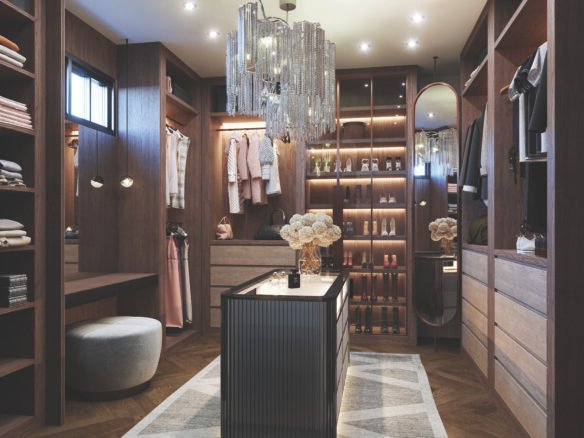 A luxurious walk-in closet with wooden shelves filled with clothes and shoes, perfect for new homes. A central island is adorned with decorative flowers under a glamorous crystal chandelier. Soft lighting creates a warm ambiance, highlighting elegant décor and design.