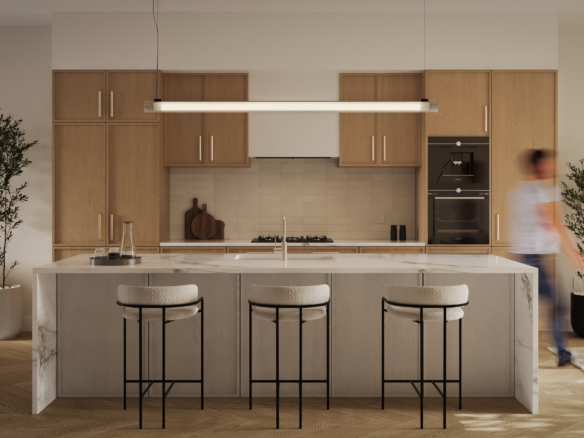 A modern kitchen in a new condo with a marble island and three cushioned barstools. Light wood cabinets complement the linear pendant light above. Stainless steel appliances and minimal decor, including plants, complete the space while a person appears slightly blurred in motion.