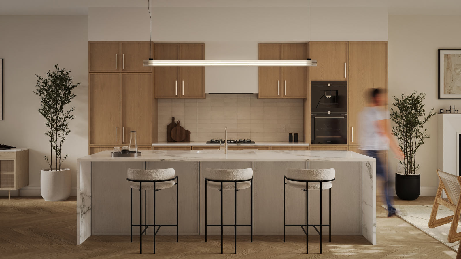 A modern kitchen in a new condo with a marble island and three cushioned barstools. Light wood cabinets complement the linear pendant light above. Stainless steel appliances and minimal decor, including plants, complete the space while a person appears slightly blurred in motion.