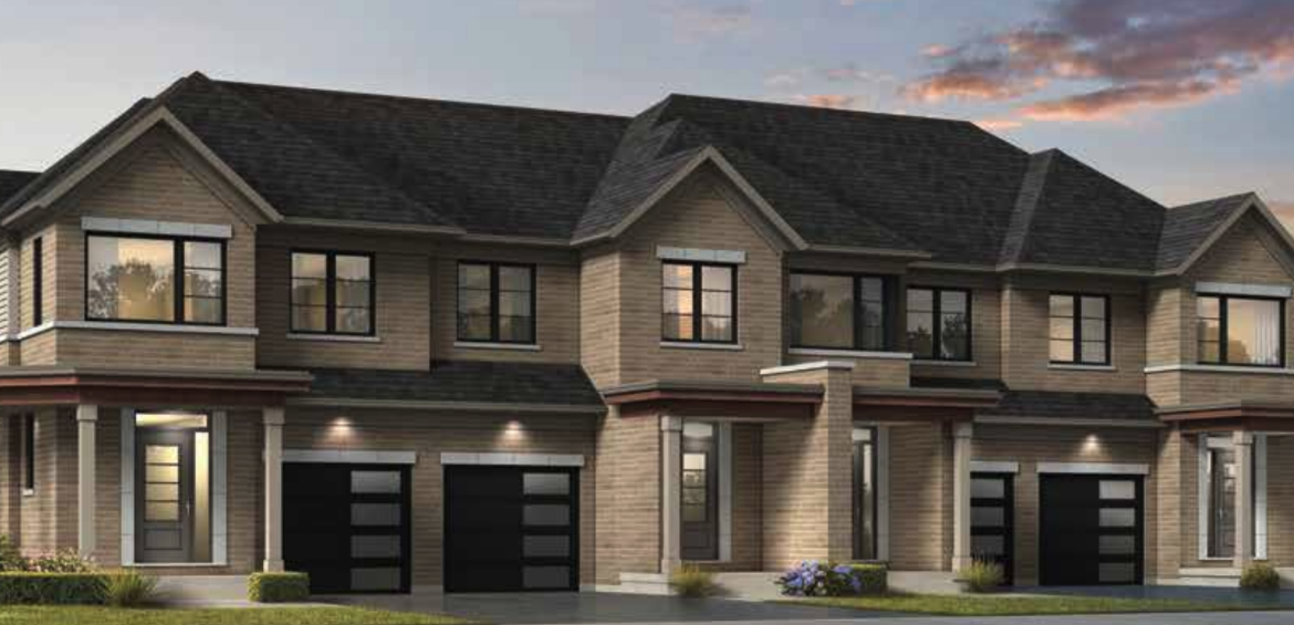 A modern townhouse complex at dusk, featuring neutral-toned brickwork, multi-pane windows, and dark garage doors. These new homes in Brampton have units with small shrubs and lawns in front, under a partly cloudy sky.