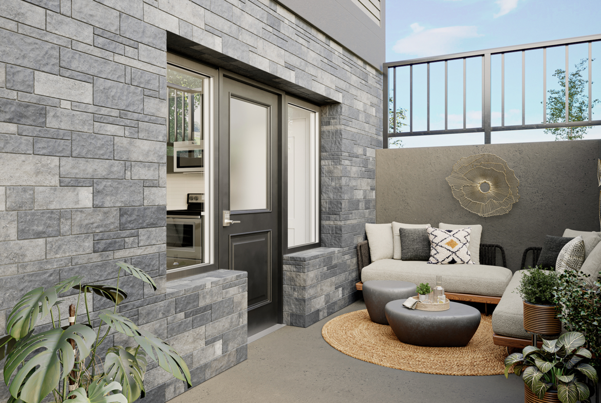 A cozy outdoor patio with a gray brick wall, a beige sectional couch adorned with patterned pillows, and a modern touch from the round woven rug and small table. The area, featuring potted plants and a tall railing, offers a tranquil and stylish Marquis setting.