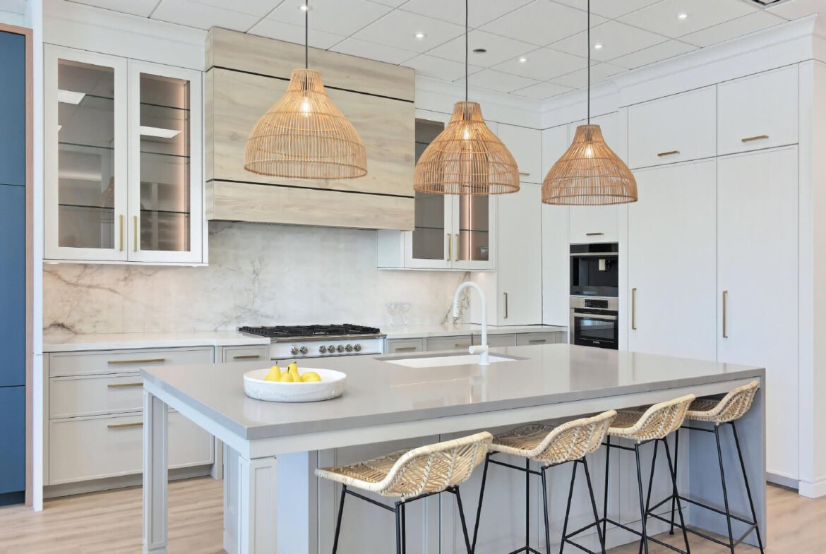Modern kitchen with a large gray island, featuring three rattan pendant lights and four wicker stools. White cabinets and a marble backsplash create a stunning backdrop. On the island countertop sits a bowl of bananas, perfect for those exploring new homes or pre-construction options in the GTA.